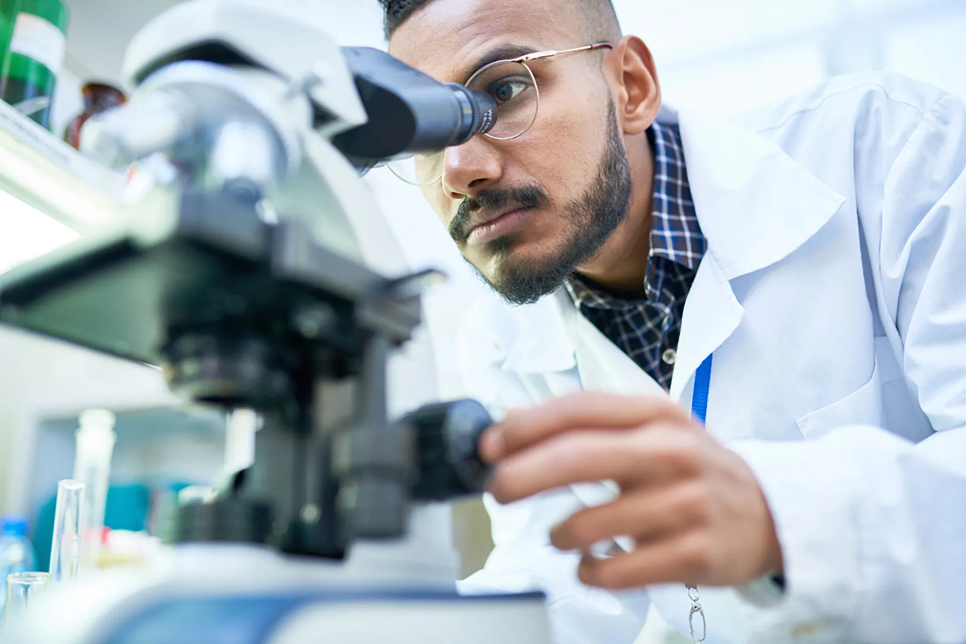 A scientist with a beard and glasses in a lab coat examining a sample under a microscope.