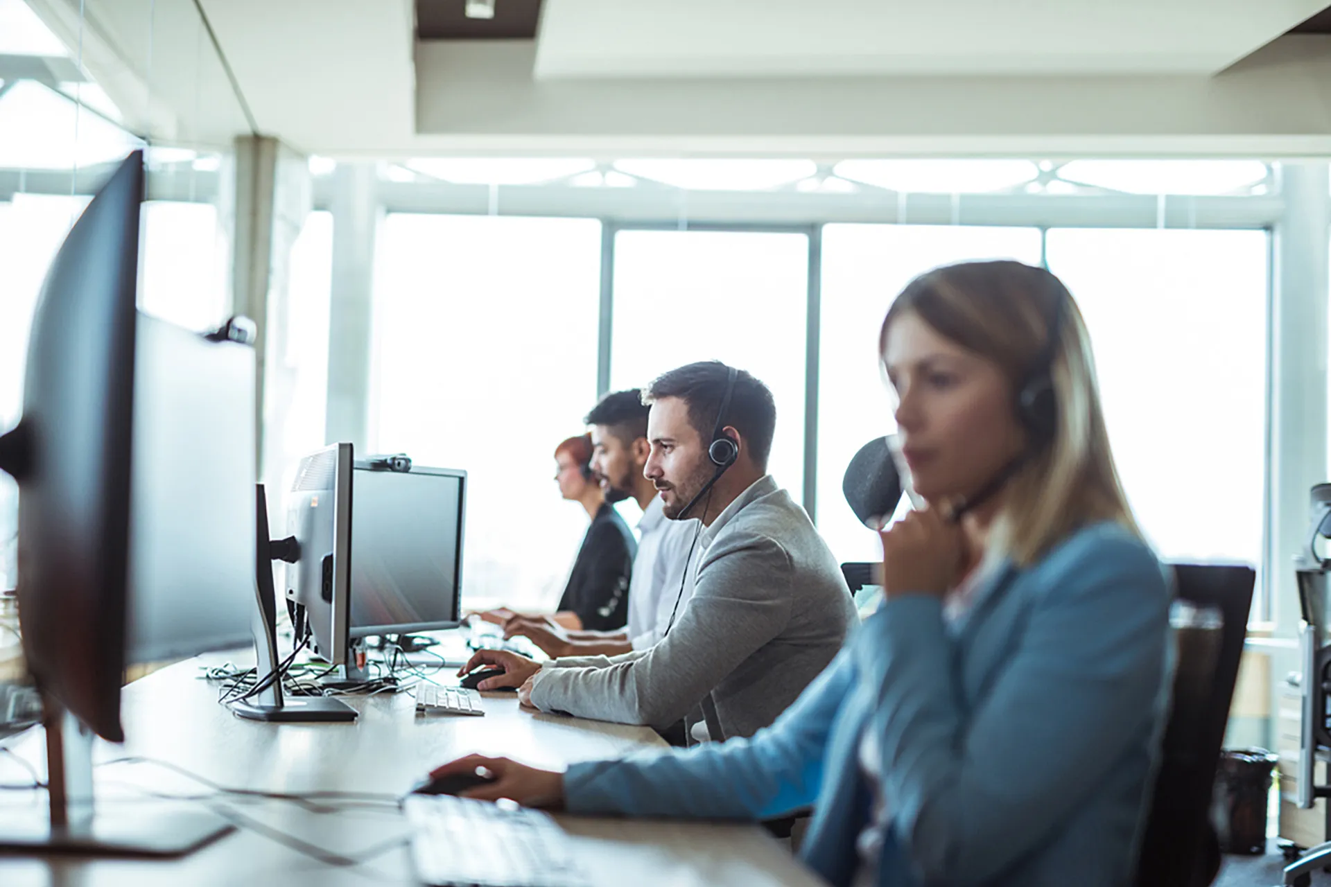 A sales team wearing headsets, working at their computers on a row of desks in a bright office.