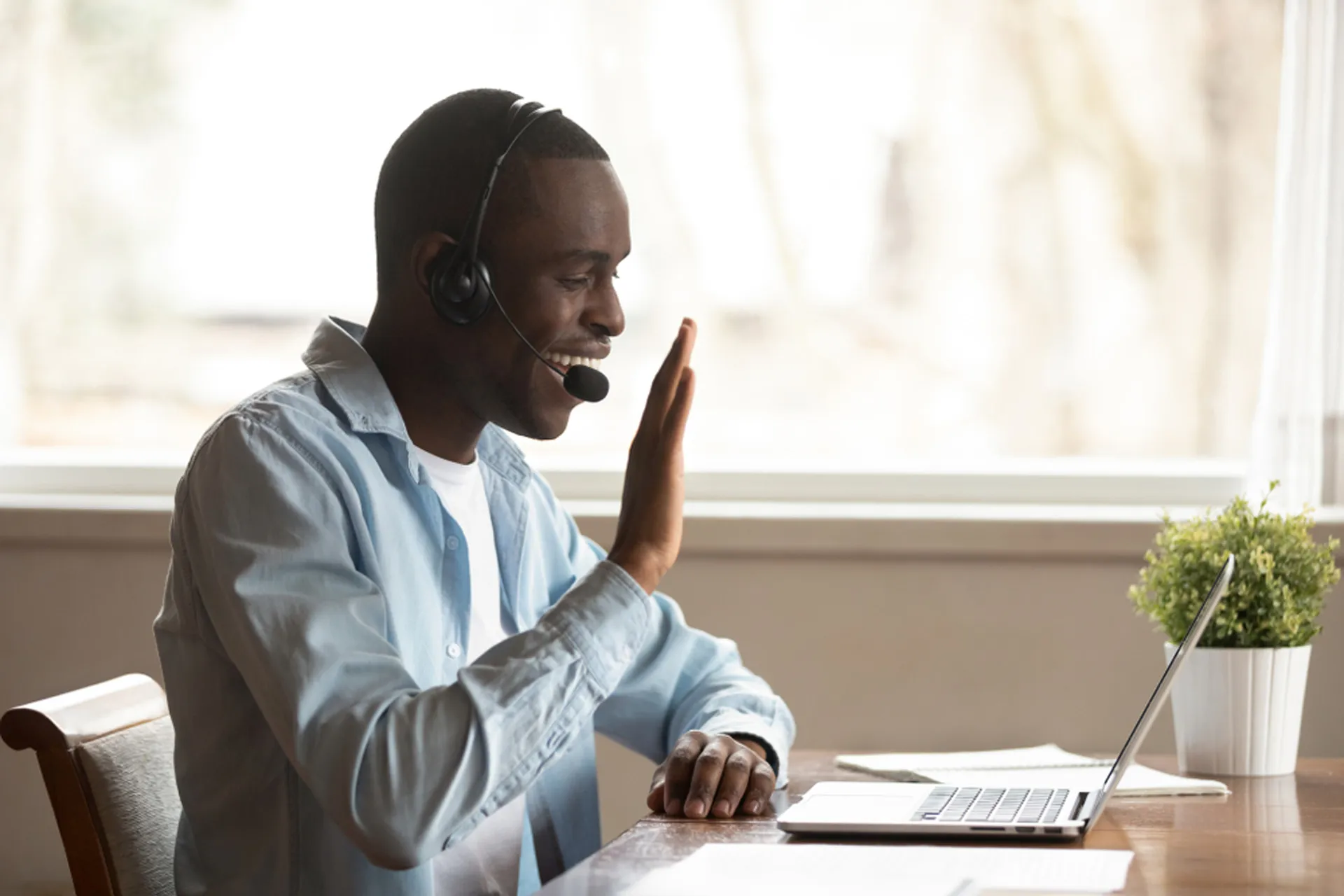 A young black man sat at his desk whilst wearing a headset, waving to someone on a video call.