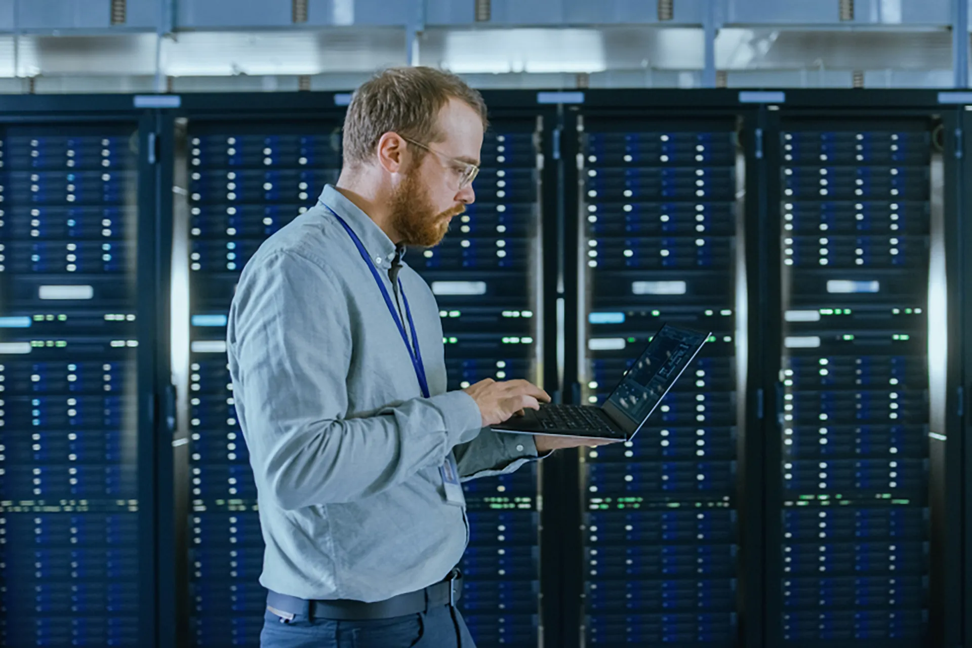 A man walks in front of a server rack, carrying a laptop.