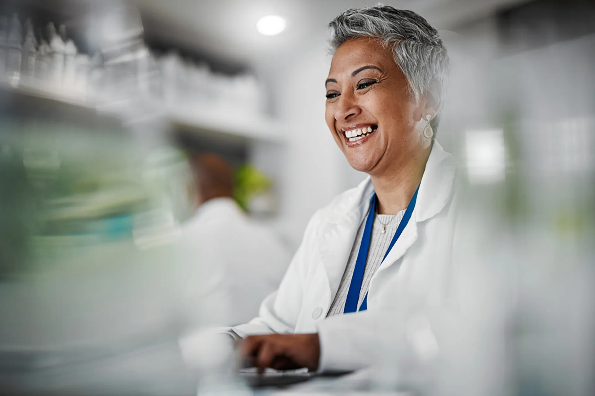 A female scientist in a lab coat smiles as she diligently works on a computer, focused and engaged in her research.