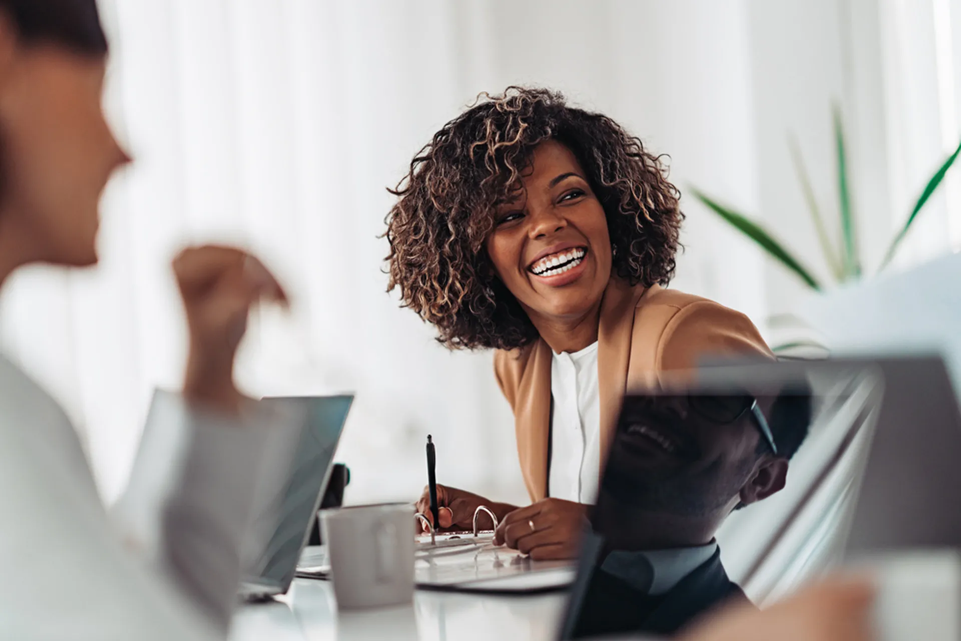 A black woman with short curly hair smiles while conversing with colleagues at a table in an office.